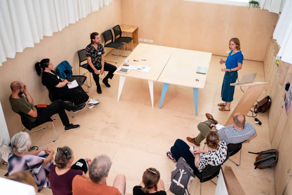Group of adults in a meeting room seated in a semicircle around a wooden table as a woman in blue explains with a laptop nearby