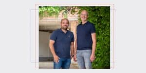 Website banners 35 - MONDEA Two men posing outdoors in front of greenery and a brick wall, wearing navy polo shirts and smiling. (informative)