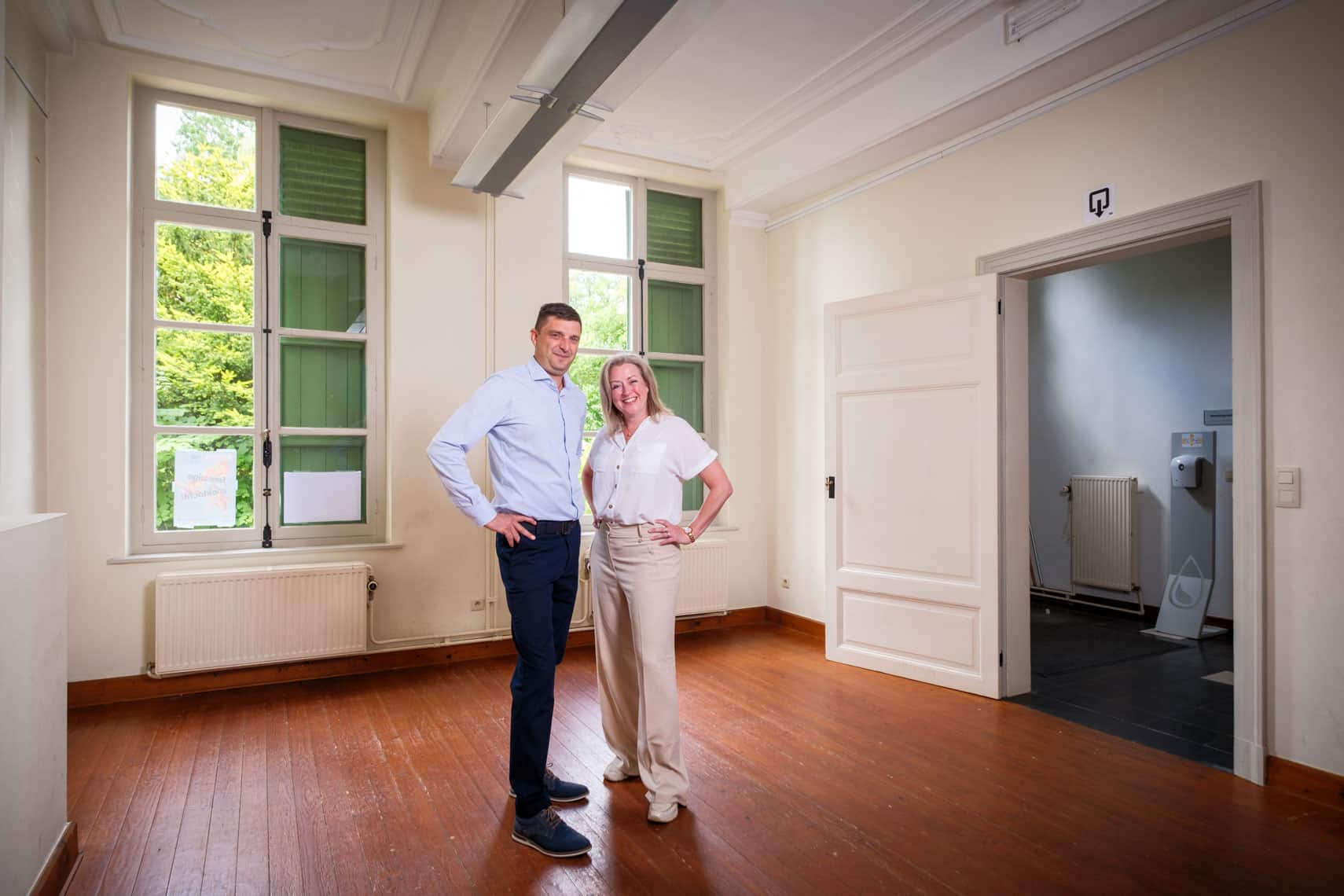 Two adults, a man in a light blue shirt and a woman in a white blouse, stand with hands on hips in a bright, empty room with large windows and green shutters.